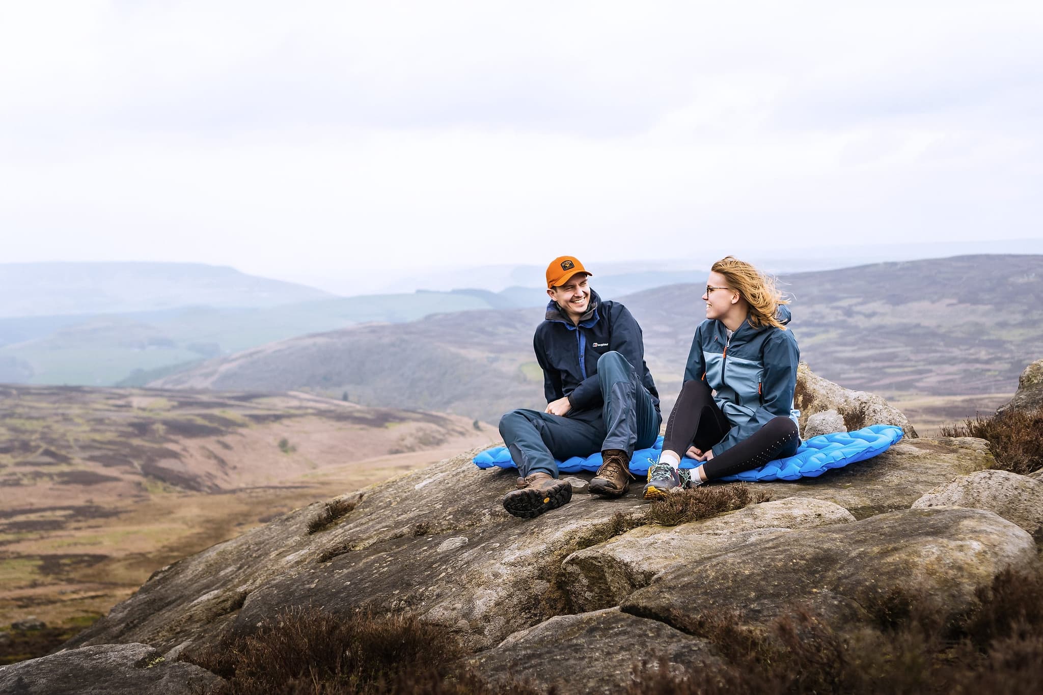 Two ramblers sat on a Rambler Peak sleeping mat on a gritstone edge above a misty Peak District valley